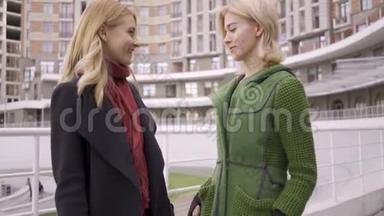 两个可爱的金发女朋友站在街上互相展示购物袋。 两个时装女人炫耀她们的